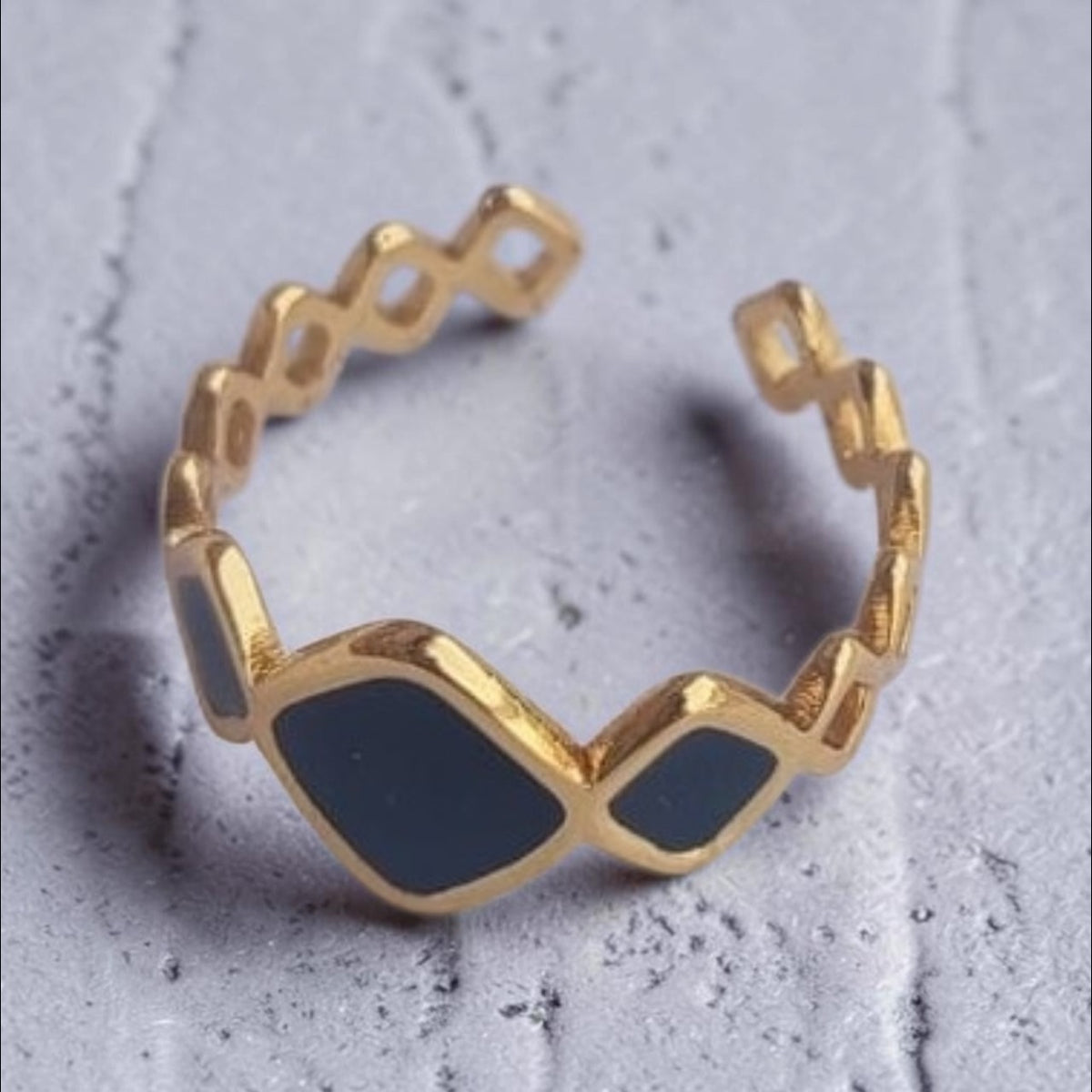 Altin Noor geometric open band ring in gold-tone metal with dark grey enamel accents on a textured grey surface.