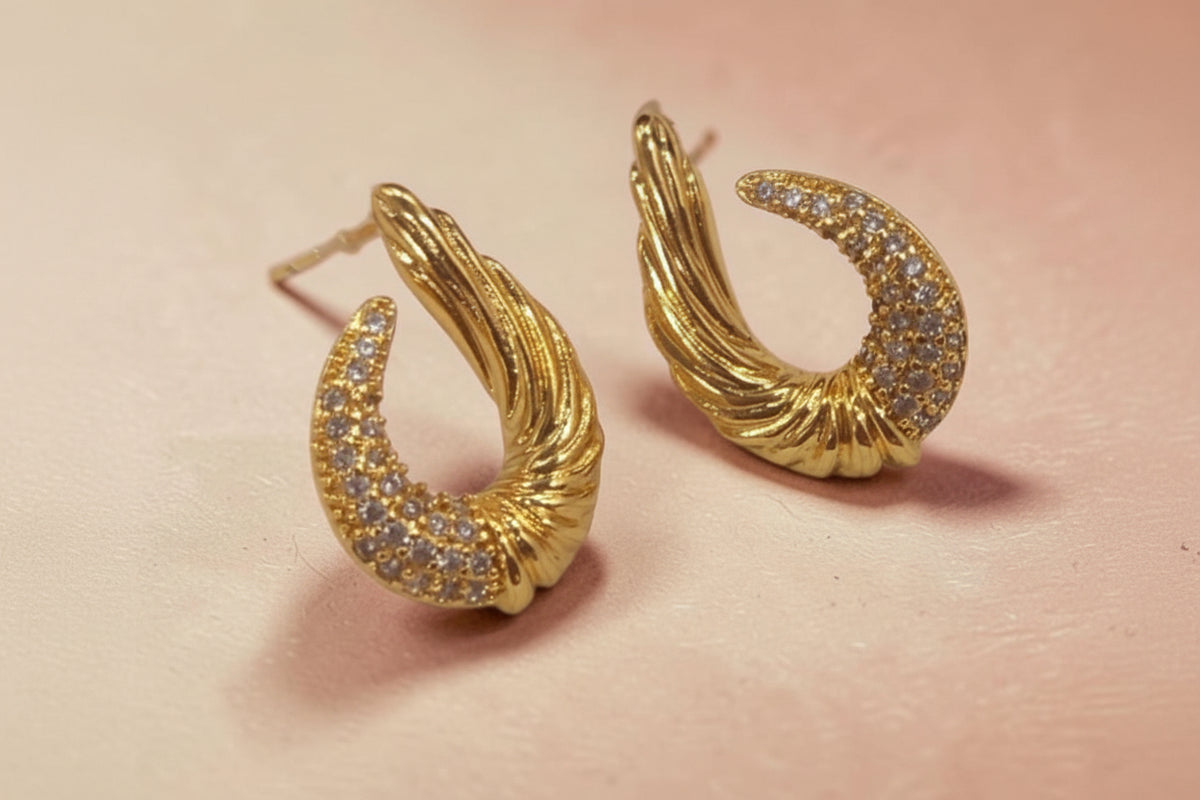 Altin Noor gold-tone crescent stud earrings with twisted metal texture and pavé-set crystals on a soft pink background
