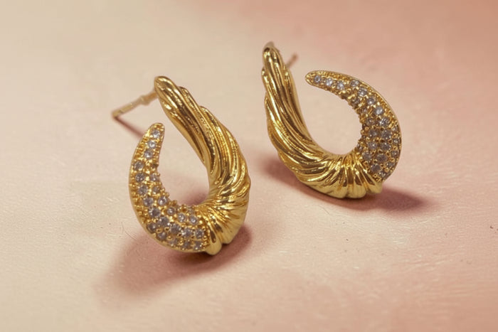Altin Noor gold-tone crescent stud earrings with twisted metal texture and pavé-set crystals on a soft pink background