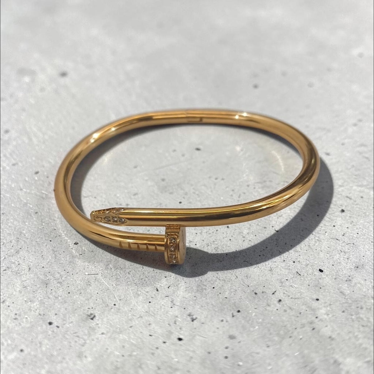 Altin Noor gold-tone nail-inspired bangle bracelet with crystal accents on a textured grey surface.